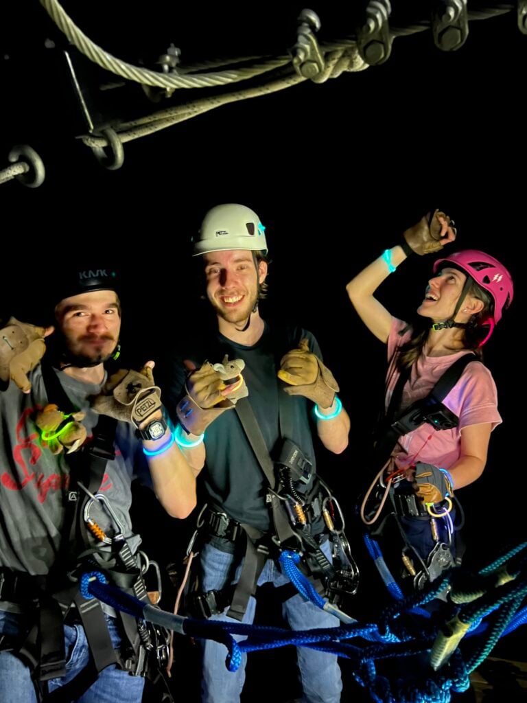 Three guests in helmets and harnesses grinning and cheering on a platform at night