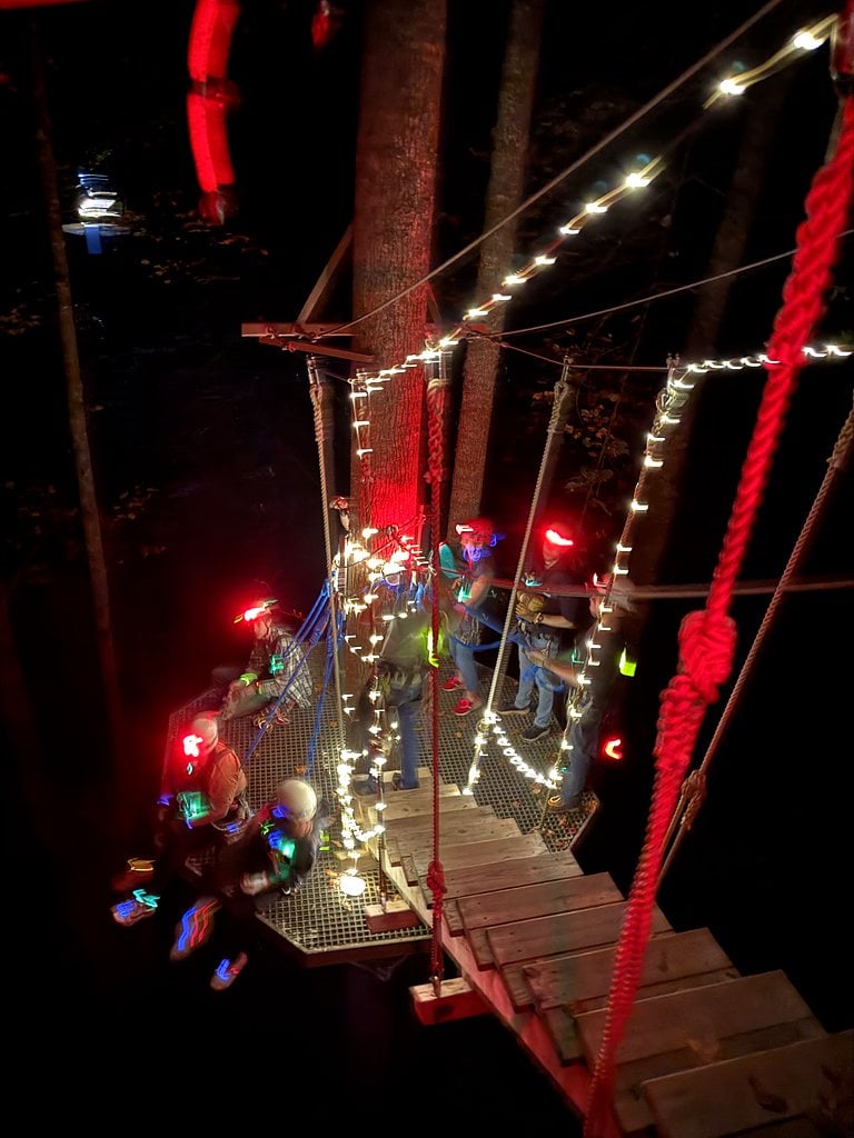 A group crossing a bridge strung with lights in the trees